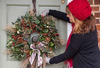 Sheree King hanging a traditional christmas wreath on a front door. Featuring Eucalyptus gunnii, Rosa 'Bonica' rose hips, Variegated Ivy, Ilex aquifolium, Cotoneaster lacteus, Pinus nobilis, Godetia and Miscanthus sinensis seed heads