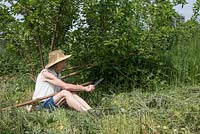 Lady cutting grass on allotment with scythe, sitting sharpening scythe blade