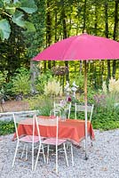 Vintage chairs and table with glass jar, vase and a pink parasol.  In the background, two stone doves on pillars. Border with ornamental grasses, Cortaderia selloana