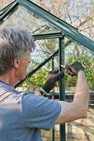 Man using needle nose pliers to insert 'w' shaped stainless steel glazing clip, to hold pane of glass in place: April, Spring.