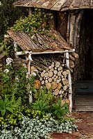 Rustic covered log store and woodland planting including Viburnum with hut behind - The Woodcutter's Garden - RHS Malvern Spring Show 2016. Designer: Mark Walker, Sponsor: Howards Motors