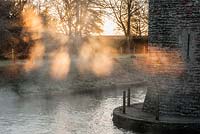 Early morning sun illuminates mist rising from spring water tumbling into the moat around the Bishop's Palace in Wells on a November morning 