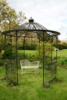Elegant circular gazebo in black painted steel set in grass with a lovers seat beneath and recently planted climbers. 