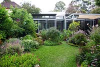 View looking from the back of garden towards rear of house, showing curved garden beds, a soft leaf buffalo lawn and a variety of flowering and colourful herbaceous plants.