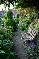 A path in The Bear Shop Gardens with a bench and mixed bed borders of Taxus baccata, Roses, Fuchsia Magellanica hybrid and Campanula poscharskyana