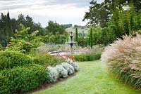 A curved grass path, bordered by clipped Cistus x loretii, Helichrysum italicum and Rosa 'Kadora' on the left and Miscanthus sinensis 'Autumn Light' on the right, leads to the Italian garden with a Florentine fountain in les Jardins de la Poterie Hillen.