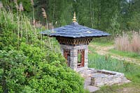 A miniature version of the Bhutan Temple. This is part of the wild area, featuring Rosa rugosa and reeds - Phragmites australis, with a grove of silver birch - Betula pendula behind.