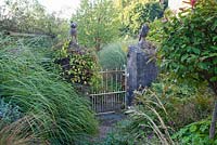 Aged metal gate with pathway amongst overgrown planting. Barn House, Gloucestershire