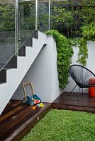 A childs play area under a set of stairs with frameless glass screens a timber deck, timber children's toy and Aptenia cordifolia, heartleaf iceplant spilling over a grey painted cement rendered wall.
