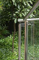 Detail of a stainless steel hand rail and support columns for glass panels on a set of stairs in front of a garden bed with a Elaeocarpus 'Luscious'.