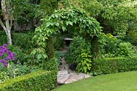 An arch covered with Virginia creeper, Parthenocissus quinquefolia, above a stone path leading to a secluded bench.