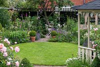 A circular lawn is enclosed in cottage style herbaceous borders, leading to shady seating, a compost bin amongst foxgloves, and greenhouse.