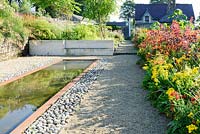 Reflecting pool edged with rusted steel and inset pebbles beside hot beds full of vivid red, orange and yellow herbaceous perennials and shrubs including yellow Leontodon rigens, orange Lilium lancifolium, Cotinus 'Grace', Dahlia australis and Crocosmia 'Lucifer'.
