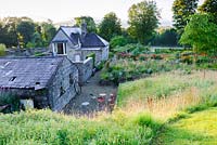 View down from hillside above the garden showing layout of former steward's house, barns, wild meadow areas, raised ornamental beds and fields and countryside beyond.