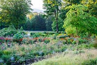Beds planted with a matrix of herbaceous perennials and shrubs set between grassy slopes in the higher part of the garden in the foreground and cultivated farmland beyond with the Wicklow mountains glimpsed through a gap in the trees.