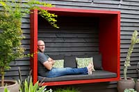 Garden owner and designer Steven Wells, relaxing in a red painted timber seating pod he built, attached to a black painted timber wall.