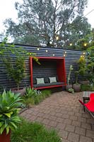 Paved area featuring a black painted timber wall with a red sitting pod attached to it and a collection of large pots with a maple and various succulents. A border planting of Australian native grass, Lomandra 'Tanika'.