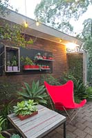 A seating area at night at the back of brick house with a bespoke recycled timber table and a retro style red canvas butterfly chair. A collection of mauve and red ceramic pots mounted on a black timber panel planted out with cactus and succulents.