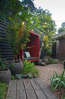 A timber walkway leading to a paved area featuring a black painted timber wall with a red seating pod attached to it. A collection of large pots with a maple and various succulents, a border planting of Australian native grass, Lomandra 'Tanika'.