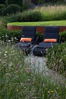 Modern rattan sun loungers on oak decking surrounded by meadow borders, Pinus mugo hedge in front of rusted Corten steel walls. 