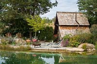 There is a wide rest area with fire bowl and deck chair next to two-storey witches' cottage. In the foreground, the natural swimming pond. Plants include Pinus sylvestris, Styrax japonica and Lespedeza thunbergii