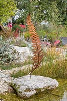 Next to the shallow water zone a metal sculptural fern on a granite boulder. In the background a rest area, birches and perennial planting