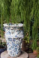 Large urn faced with pieces of broken blue and white china surrounded by a weeping juniper. Sculptor and ceramicist Marcia Donahue's garden in Berkeley, California.