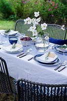 Outdoor dining table dressed in shades of blue and a vase of white Japanese anemones.  Pockets are made in the folded napkins to put name card and each setting is finished with an olive branch