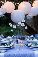 Outdoor dining table dressed in shades of blue and with a vase of white Japanese anemones.  Paper ball lanterns, fairy lights and paper pompoms hang over the table to decorate. Pockets are made in the folded napkins to put name card and each setting is finished with an olive branch.  Tealights are put on the table to light it up as dusk falls