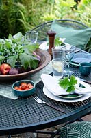 Outdoor dining area under a fig tree on a brick patio.  Table laid with greens and fresh whites, napkins dressed with a geranium leaf and the centre of the table has a wooden bowl of fresh green leaves from the garden surrounded by fruit