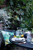 Dusk falls on the outdoor dining area under a fig tree. Fairy lights are in the trees creating a cosy setting and light up the area. Table laid with greens and fresh whites and napkins dressed with a geranium leaf