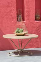 Beneath A Mexican Sky Garden - View of patio table with a terrarium with succulent, rendered red wall with alcoves and plants growing in glass vases - RHS Chelsea Flower Show 2017