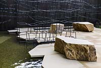 Thin black metal rods sculpture of 'Disappearing Walls' and angular sandstone blocs on fragmented large scale paving overhanging reflective pool - Breaking Ground - RHS Chelsea Flower Show 2017 - Designers: Andrew Wilson and Gavin McWilliam - Sponsor: Darwin Property Investment Management Ltd