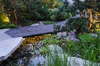 Wooden path leading to the terrace made from sliced boulders with abstracted paving design and illuminated pond surrounded by  Pinus banksiana - Jack pine and Iris x robusta 'Gerald Darby' - The Royal Bank of Canada Garden - RHS Chelsea Flower Show 2017 - Designer: Charlotte Harris - Sponsor: RBC
