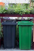 The RHS Greening Grey Britain Garden - Wheelie bins for household rubbish and green wheelie bin for recycling with a living roof cover planted with edible plants and herbs, viola, chives, strawberries, thyme and mint - RHS Chelsea Flower Show 2017
