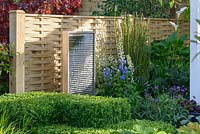 A cascade water feature in front of fence surrounded by  clipped box hedge and planting of Zantedeschia, Verbena, Kale, blue Delphinium and grass - Calamagrostis x acutiflora 'Karl Foerster' - BBC Gardener's World Live, Birmingham 2017 -The Anniversary Garden: A Brief History of Modern Gardens - Designer: Prof. David Stevens