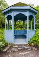 Blue painted wooden gazebo with built-in sitting benches and cedar shingles covered in green Bryophyta - Moss overlooking pond in summer, Centre de la Nature public garden, Saint-Vincent-de-Paul, Laval, Quebec, Canada