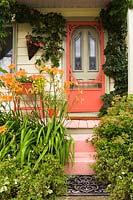 Old cottage style house facade with red door and veranda with climbing Hedera helix. Border with Hemerocallis and Spiraea shrubs.