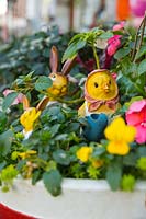 A pot with yellow Violas, pansies, pink Impatiens, two rabbits and a chick with yellow faces and decorations in the pot.