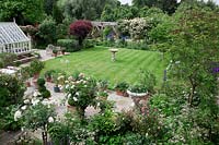 Overview of garden with greenhouse and cold frames, neat stripy lawn, paved terrace with pots and Lutyens bench, oak pergola. Planting includes Cotinus coggyria, Rosa 'Dublin Bay' ,Rosa 'Crocus', Rosa 'Iceberg', Rosa 'Phyllis Bide', Rosa 'Long John Silver'