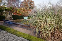 Miscanthus sinensis 'Variegatus' planted by Buxus - Box - low hedge containing Lavender. Blue painted bench by Water Lily filled pond,  Phormium tenax purpureum group planted in old copper tub backed with Vitis coignetiae.
