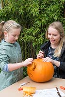 A young girl removes the cutout section of an eye on a large pumpkin, helped by her mother