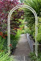 A metal arch straddles a hidden side path, beside red leaved American redbud tree and helenium.
