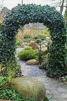 Arch of Hedera (ivy) over pathway leading to a garden of shrubs large boulders in John Massey's garden at Ashwood Nurseries