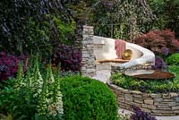 The Kingston Maurward Garden at RHS Chelsea Flower Show 2019. A curving seating area in Purbeck stone, surrounded by purple beeches, acers and other herbaceous planting. Design: Michelle Brown. Sponsors: Miles Brown, Kingston Maurward College, Goulds Garden Centre, Wilks Landscaping, Holme for Gardens, The Green Gardener.