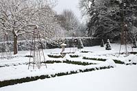 View of snow-covered, formal garden, featuring low Buxus sempervirens - Box - beds with decorative iron obelisks, a central sundial and Taxus baccata - Yew - cones.
