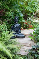 A black Buddha statue sits on a path built with railway sleepers amongst the lush foliage planting. 