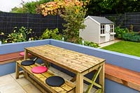 Wooden table and bench dining area look out to child's playhouse in modern garden. 