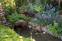 Stone arch over a stream next to vegetable garden with leeks, cabbage, chives, lettuce and chard - Welcome to Yorkshire Garden - Sponsor: Welcome to Yorkshire - RHS Chelsea Flower Show 2018