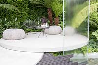 VTB Capital Garden - Spirit of Cornwall - Concrete patio area with wooden seats, table, chair with glass water wall and Dicksonia antarctica - Sponsor: VTB Capital - RHS Chelsea Flower Show, 2018